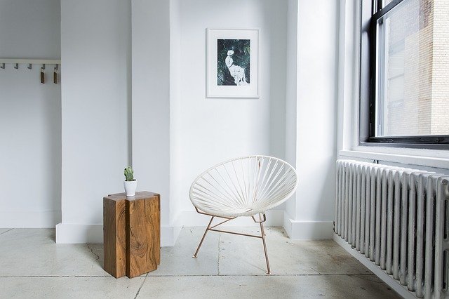 Weißer Raum mit Baumstamm als Tisch, einem weißen Stuhl, einem Bild an der Wand und links ein Fenster und darunter eine Heizung.
