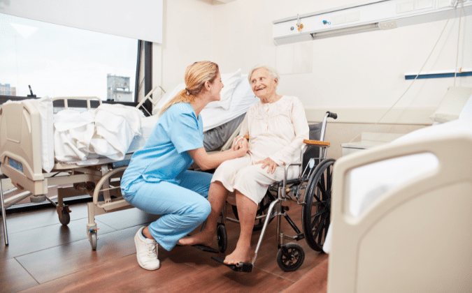 Pflegerin hockt vor einer älteren Dame im Rollstuhl und hält ihre Hand