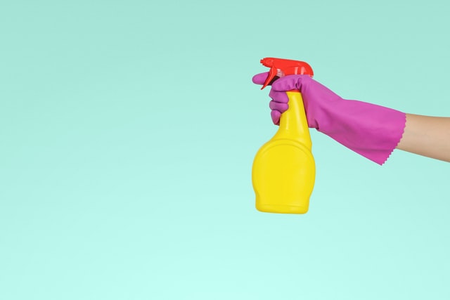 Hygienewaschmittel: sinnvoll, notwendig oder überflüssig? - Das Foto hat einen hellblauen Hintergrund. Man sieht eine Hand mit einem lila Reinigungshandschuh und einer gelben Sprühflasche mit rotem Sprühhals.